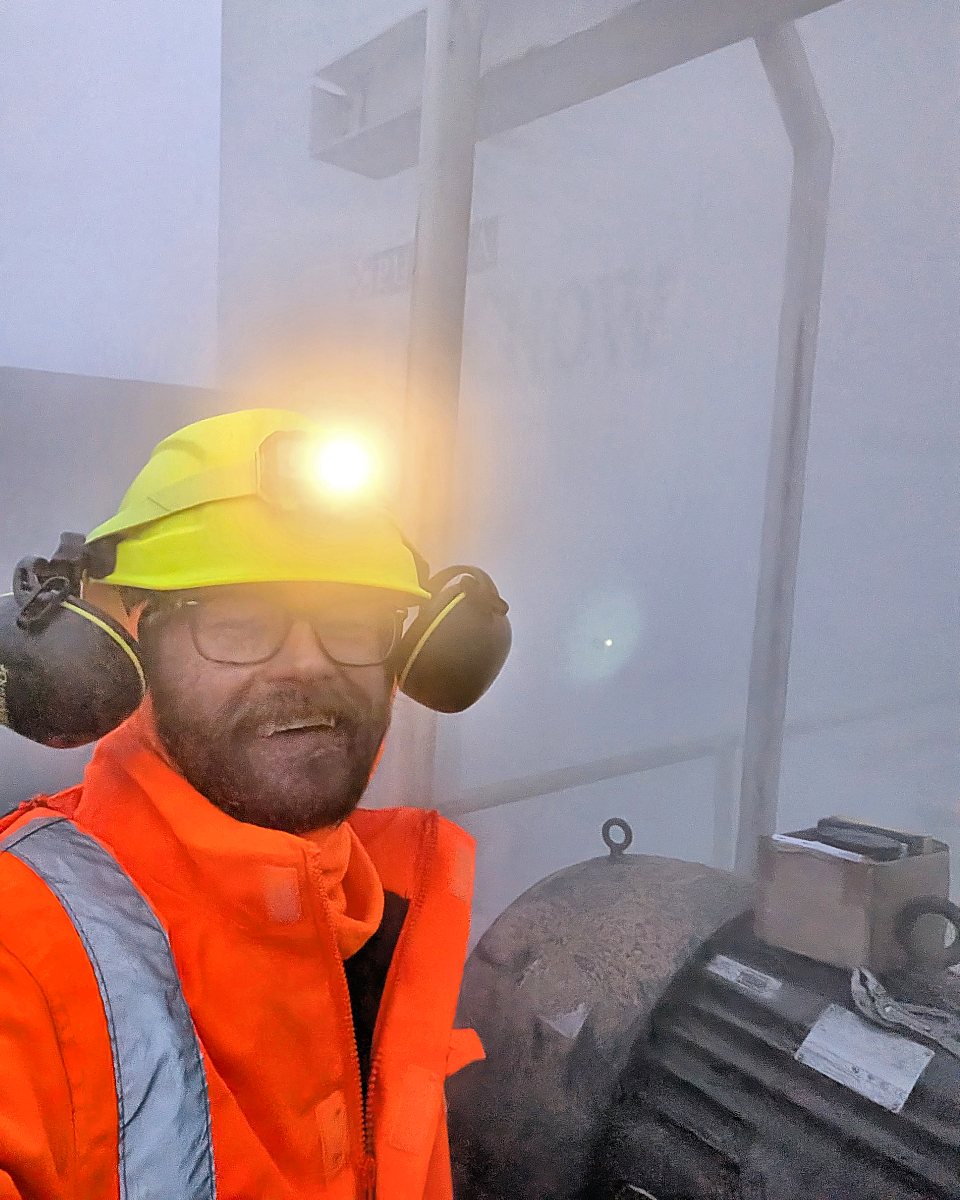 Michael McKenzie in a hard hat, headlamp, hearing protection and hi-vis jacket, next to an industrial motor on a foggy morning